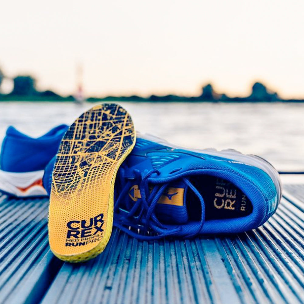 Photograph of a Currex insole leaning against a blue running shoe on a wooden surface near a lake or river
