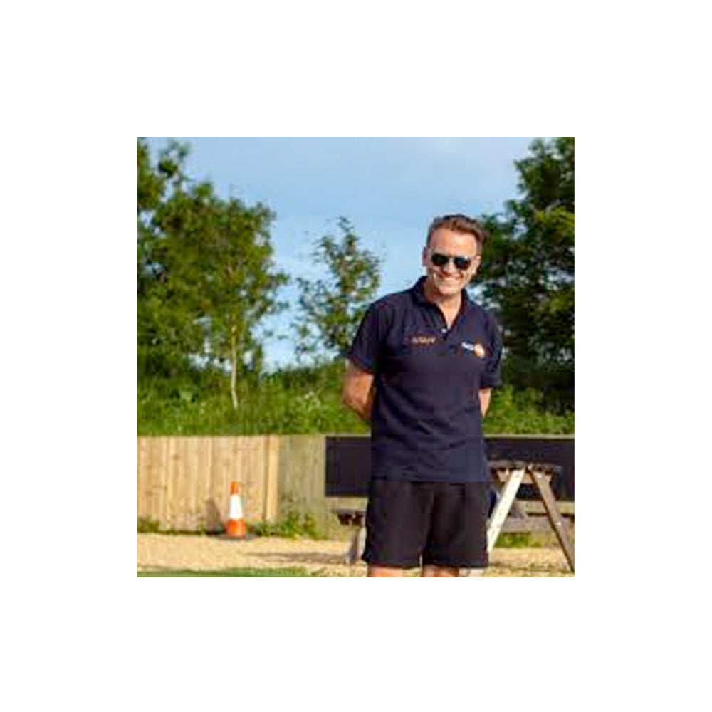 Man wearing a navy blue polo shirt with a logo, standing outdoors with trees and a fence in the background.