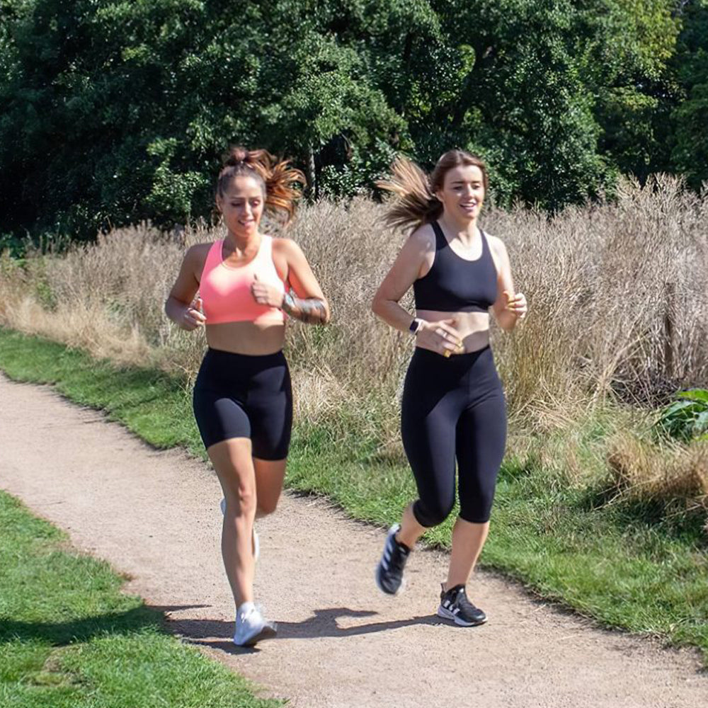 Photograph of two women running on a path in a park with trees and grass in the background.