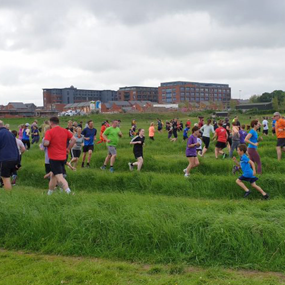 Runners in a race on a grassy field at Somedale Pavilion Parkrun with buildings in the background