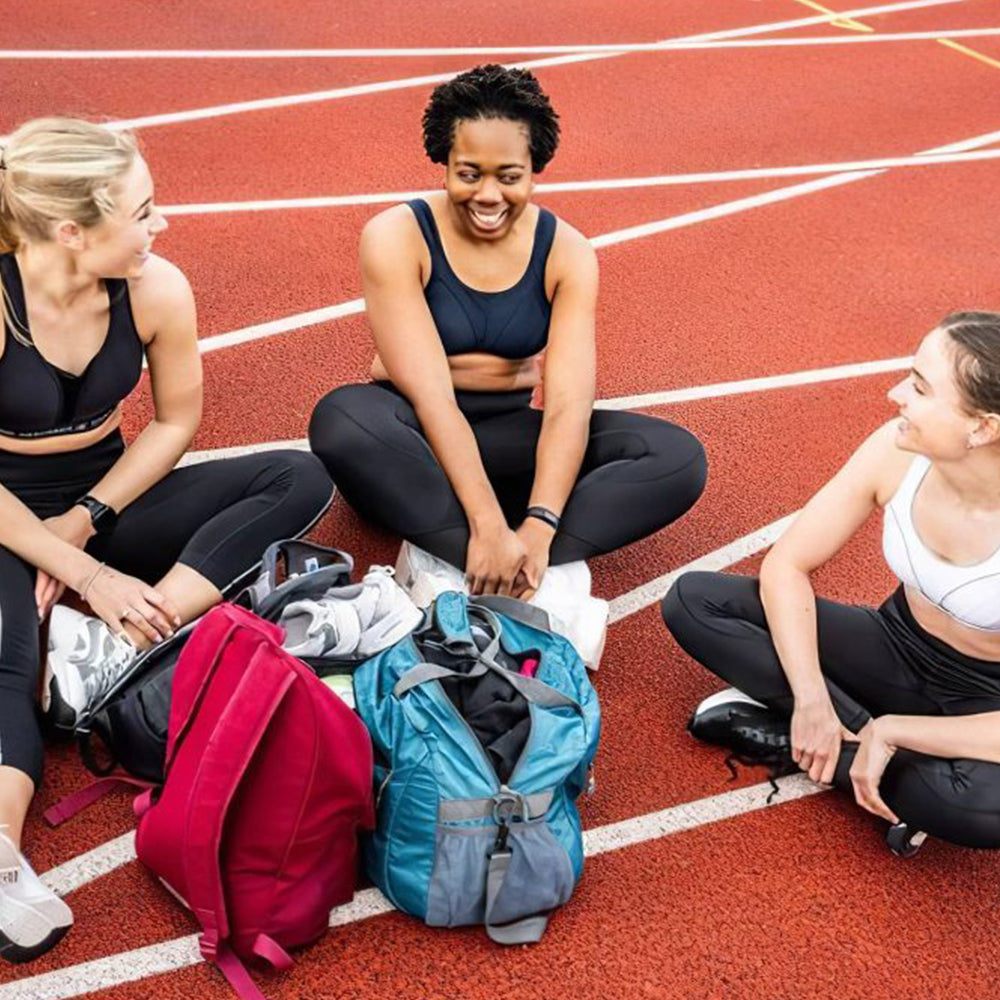 Photograph of three women wearing running bras and running tights sitting on a running track with sports bags around them